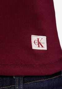 Tissu côtelé bordeaux avec une étiquette blanche Calvin Klein portant un texte rouge et un logo 'CK' distinctif près de l'ourlet, associé à un jean bleu.