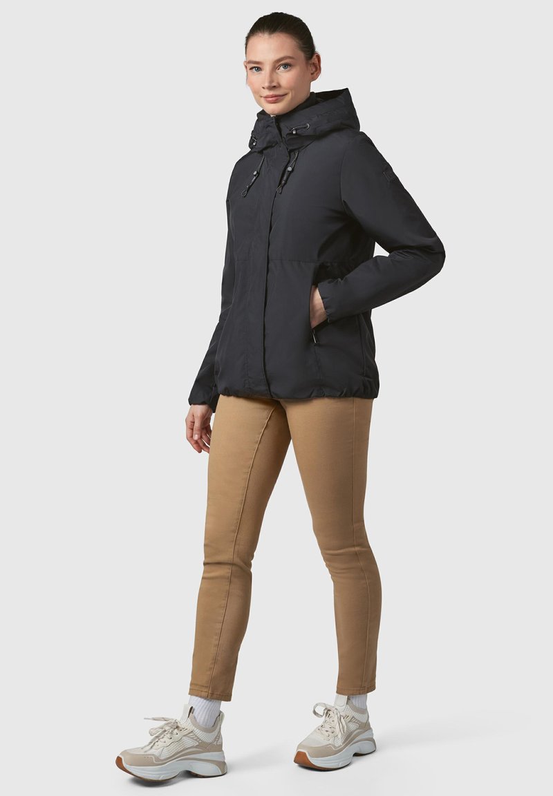 Črna kapuca jakna z zadrgo spredaj, elastičnimi manšetami in stranskimi žepi, v kombinaciji s peščenimi hlačami in bež superge. Eleganten in funkcionalen dizajn.