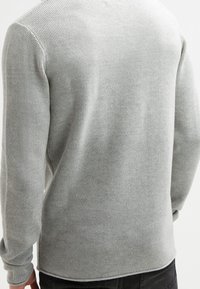 Světle šedý texturovaný pletený svetr nošený osobou z pohledu zezadu s černými kalhotami, mírně pokrčenou levou paží a viditelnými detaily šití.