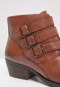 Bottine en cuir marron avec trois sangles à boucles, talon en bois empilé, texture lisse et design incurvé.