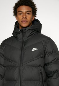 Crna puf jakna s kapuljačom, s patentnim zatvaračem sprijeda, Nike logotipom na prsima i horizontalnom prošivanom teksturom. Podesiva vezica u kapuljači.
