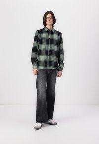 Camisa de franela en un patrón de cuadros verde y negro con botones, mangas largas y un cuello, combinada con jeans acampanados de color gris oscuro y zapatillas.