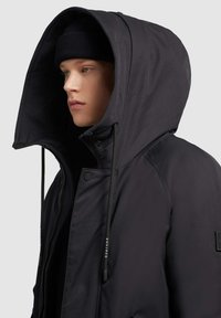 Svart huva jacka med dragkedja och tryckknappsförslutning, utrustad med en hög krage, dragsko och texturerat tyg på ärmarna.