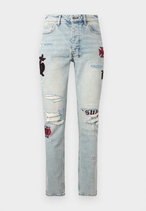 Φωτεινά γαλάζια τζιν με φθαρμένη όψη, με μπαλώματα και σκισίματα στους μηρούς και στα γόνατα, διακοσμημένα με κόκκινα και μαύρα κεντήματα.