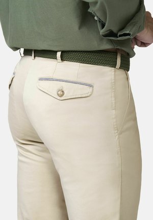 Beige broek met een achterzak met knoop en een zijnaadzak, gecombineerd met een groene geweven riem en een olijfgroen overhemd dat is ingestopt.