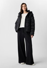 Kobieta w czarnej kurtce puchowej nosząca biały top, czarne spodnie w szerokim kroju i białe sneakersy, stojąca na tle jednolitym.