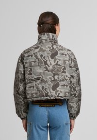 Pelēka kamuflāžas pūkains mēteļa ar augstu apkakli. Tam ir izšūts dizains un elastīgas aproces, kombinēts ar gaiši ziliem džinsiem un zīmola jostu.