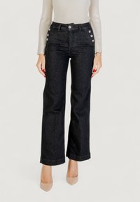 Nainen, joka on pukeutunut korkeavyötäröisiin mustiin farkkuihin, joissa on hopeiset napit taskuissa, beigen värisiin korkokenkiin ja beigen väriseen ribattuun pitkähihaiseen puseroon.