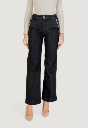 Nainen, joka on pukeutunut korkeavyötäröisiin mustiin farkkuihin, joissa on hopeiset napit taskuissa, beigen värisiin korkokenkiin ja beigen väriseen ribattuun pitkähihaiseen puseroon.