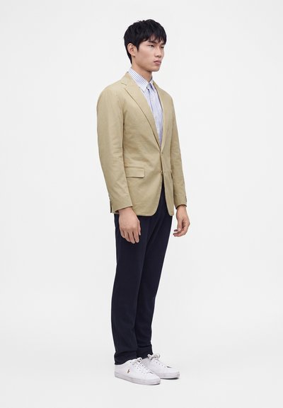 Ung mand iført beige blazer, stribet skjorte, mørke bukser og hvide sneakers foran hvid baggrund.