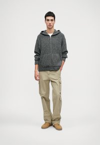 Šedá, textúrovaná pletená mikina s zipsom a prednými vreckami, skombinovaná so svetložltými cargo nohavicami so bočnými vreckami a vyhrnutými manžetami a hnedými topánkami.