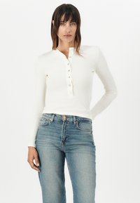 Hvit langermet topp med rund hals og knappeglidelås. Brukt med lyseblå høytlivsjeans. Slitesterk tekstur, avslappet passform.