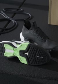 Paire de chaussures de sport noires, dont une avec une semelle verte et noire arborant le logo Continental, posée sur un sol sombre de salle de sport près d'une bande de résistance enroulée.