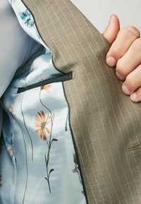 Khaki gestreifter Jackeninneres mit blauer floraler Satin-Fütterung. Verfügt über eine kleine Tasche mit schwarzem Rand und detaillierten Blumenmustern.