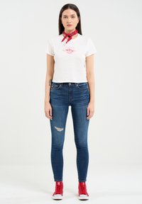 Balts īss krekls ar sarkano grafiku, apvienots ar tumši ziliem šaurajiem džinsiem ar izsistām ceļgala vietām, un sarkaniem augstiem sporta apaviem.