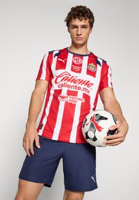Rød og hvit stripete fotballdrakt med blå krage, kombinert med marineblå shorts. Spilleren holder en svart og hvit fotball med en rød logo.
