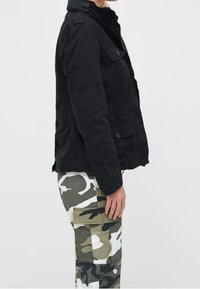 Giacca nera con texture, colletto alto e tasche frontali, abbinata a pantaloni cargo camouflage verde e bianco con grandi tasche laterali.