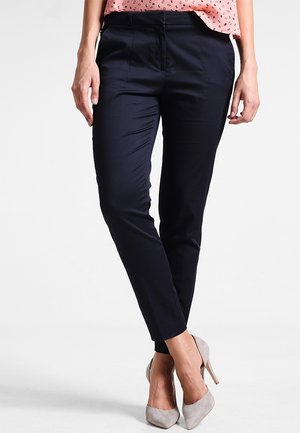 Femme portant un pantalon noir slim aux chevilles et des chaussures à talons gris, debout jambes croisées sur fond blanc.