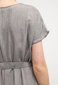 Harmaa, lyhythihainen vaate, jossa on pehmeä, mokkanahkamainen rakenne. Sisältää sidottavan vyötärön ja rentoa laskeutumista takana.