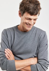Maglione grigio a maniche lunghe con polsini e collo a coste, realizzato in materiale morbido; presenta un piccolo logo sulla manica sinistra.