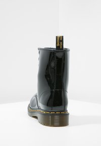 Bottine noire en cuir brillant avec surpiqûres jaunes, semelle épaisse translucide et languette noire avec texte doré à l'arrière.