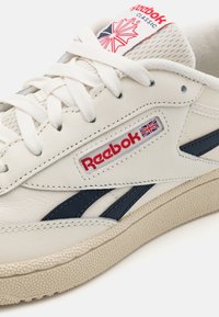 Vita lädersneakers med marinblå detaljer, med klassisk Reebok-logotyp, texturerad beige sula och platta snören. Designen inkluderar en mesh-tunga.