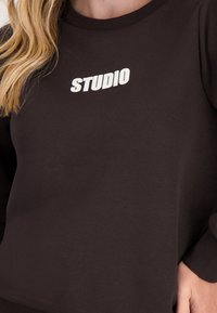Bruine sweatshirt gemaakt van zachte stof met een ribbelkraag. "STUDIO" in het wit gedrukt op de borst. Getailleerde mouwen.