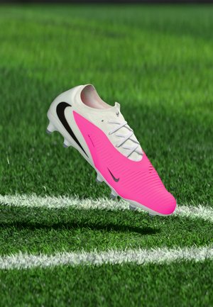 Crampon de football Nike blanc et rose avec un swoosh noir flottant au-dessus de l'herbe verte près d'une ligne blanche du terrain.