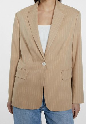 Femme portant un blazer beige à fines rayures avec un seul bouton sur un haut blanc et un jean bleu sur fond uni.