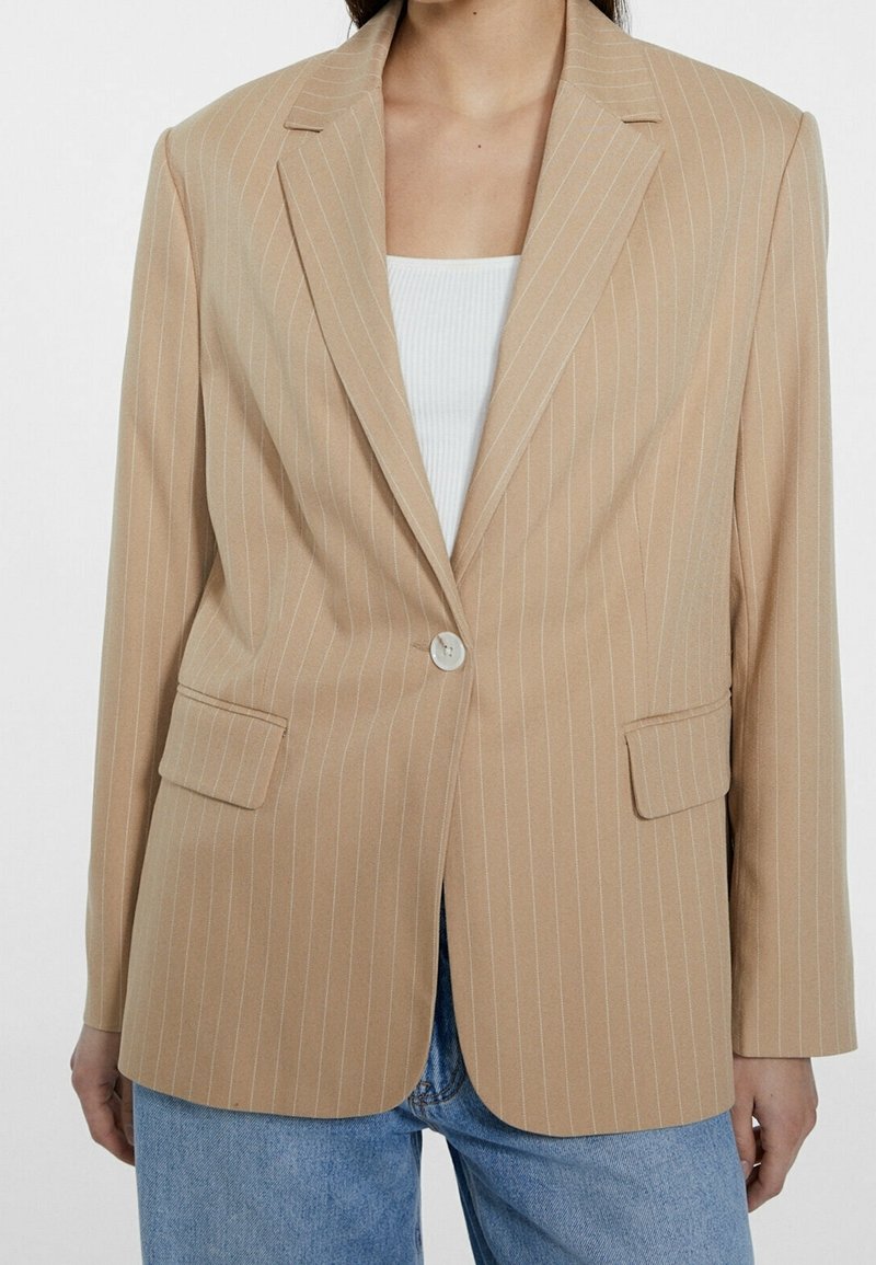 Femme portant un blazer beige à fines rayures avec un seul bouton sur un haut blanc et un jean bleu sur fond uni.