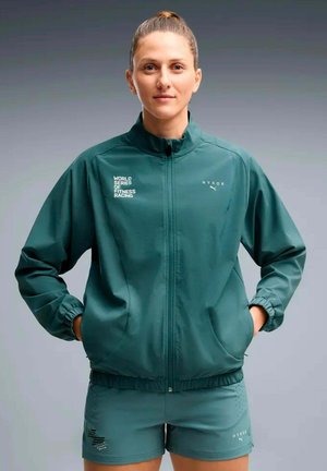 Femme aux cheveux attachés portant une veste de sport turquoise et un short avec les logos « World Series of Fitness Racing » et « HYROX », les mains dans les poches de la veste.