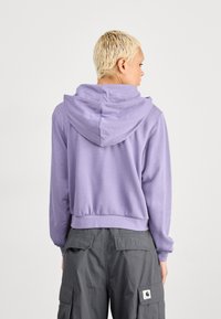 Persona con capelli corti biondi che indossa un hoodie lavanda e pantaloni cargo grigio scuro, di spalle contro uno sfondo neutro.