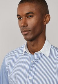 Camisa a rayas azul y blanca con un cuello blanco en contraste. La tela parece ligera y suave, con un detalle de tapeta abotonada.