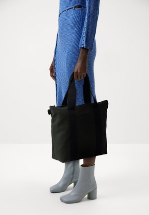 Svart tote bag med doble håndtak, glatt, slitesterkt materiale, rektangulær form og kontrasterende svarte stropper. Bærer sammen med lyseblå ankelstøvler.