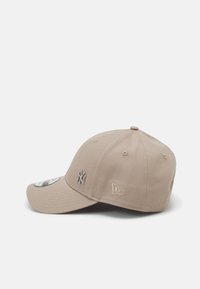 Gorra de béisbol beige con visera curvada. Presenta un logo bordado en gris y etiquetas laterales. Fabricada con un tejido texturizado y detalles de costura.