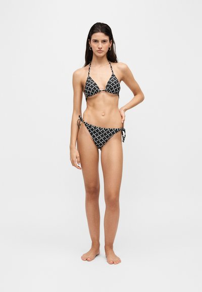 Femme debout pieds nus portant un bikini à motif noir et blanc avec un haut dos nu et un bas de bikini noué sur les côtés sur un fond clair uni.