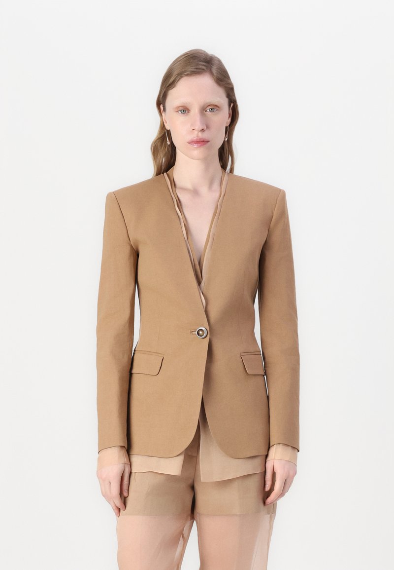 Tans kleurige blazer met een diepe halslijn en lange mouwen, voorzien van een enkele knoopsluiting en voorzakken, gecombineerd met doorschijnende shorts.