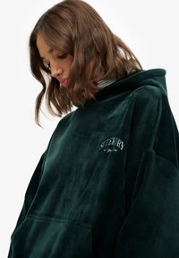 Hoodie en velours côtelé vert forêt avec logo "Superdry" brodé, capuche spacieuse et poche avant, offrant une finition douce et texturée.
