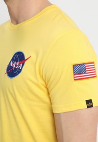 Žluté tričko s krátkým rukávem s logem NASA na hrudi a nášivkou americké vlajky na pravém rukávu, nošené osobou.