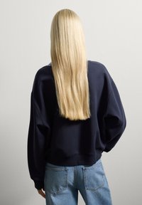 Persona rubia con el cabello largo y recto, vestida con una sudadera azul marino y jeans azules, de pie frente a una pared blanca lisa, dando la espalda al espectador.