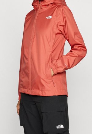 Veste à capuche corail et pantalon cargo noir avec logos "The North Face", main dans la poche de la veste, debout devant un fond clair.