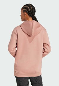 Felpa rosa con cappuccio regolabile, texture morbida, maniche lunghe e vestibilità rilassata. Indossata sopra pantaloni scuri su uno sfondo neutro.