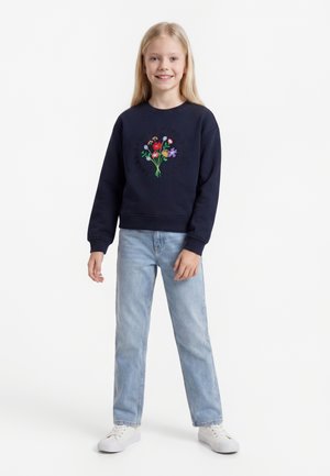 Lächelndes junges Mädchen mit langen blonden Haaren, das einen marineblauen Sweatshirt mit Blumenstickerei, hellblaue Jeans und weiße Sneaker trägt.