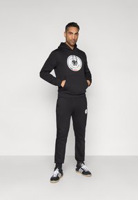 Black hoodie with front pocket, featuring a circular logo design. Paired with black jogger pants and black sneakers with white accents.