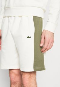 Biezi baltie šorti ar olīvzaļa sānu paneļiem. Ar zaļā krāsā izšūtu Lacoste logotipu. Mīksta kokvilnas maisījuma audums ar gludu tekstūru.