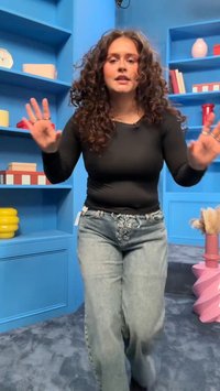 Femme aux cheveux bouclés portant un haut noir et un jean clair, levant les mains, debout dans une pièce bleue avec des étagères et des objets décoratifs.