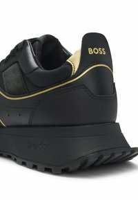 Mustat lenkkarit, joissa on teksturoitu yläosa, kultaiset yksityiskohdat ja paksu musta pohja. "BOSS"-logo on kohokuvioitu takana ja sivussa.