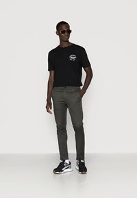 Black t-shirt with white logo, green slim-fit pants, and black sneakers with gray accents. Model wearing brown sunglasses stands casually.