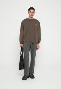 Brown sweatshirt with a subtle logo, paired with gray, fitted pants and black sneakers. The model holds a black bag. Minimalist design.