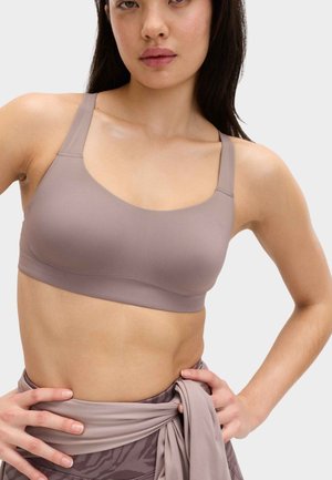 Kvinde iført en mauve sports-bh og mønstrede leggings med et bundet stofbælte, hænderne hvilende på hofterne mod en hvid baggrund.